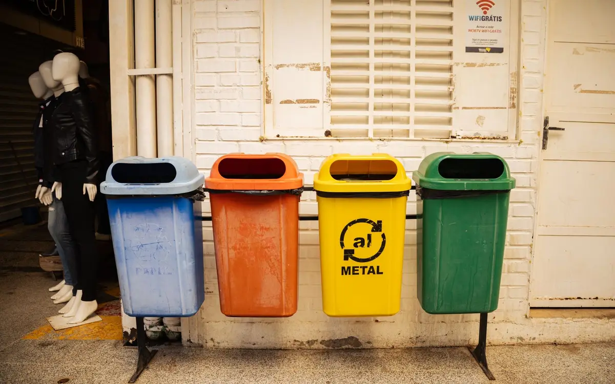 A row of color-coded commercial recycling bins for paper, plastic, metal and glass against a brick wall