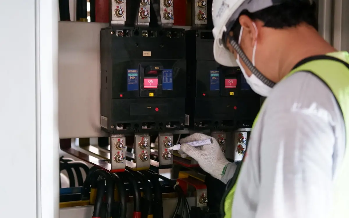 A maintenance technician in a hard hat and hi-vis vest checking breakers on an electrical distribution panel